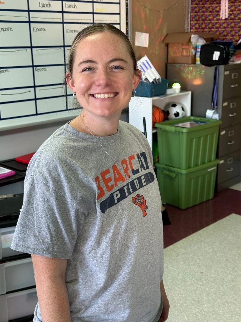 A district staff member is standing in a school classroom. A whiteboard and shelves with sports equipment and boxes are visible in the background.