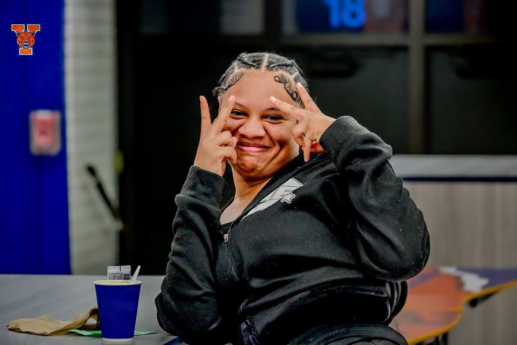 A student is sitting at a table in a school cafeteria holding up the peace sign. A cup and napkin can be seen on the table.