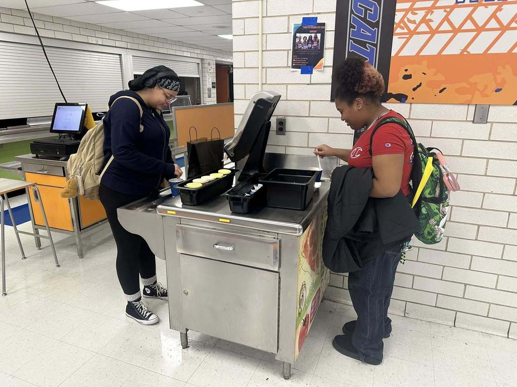 Two students are standing at a school lunch counter in a school cafeteria mixing a drink together with a plastic spoon.