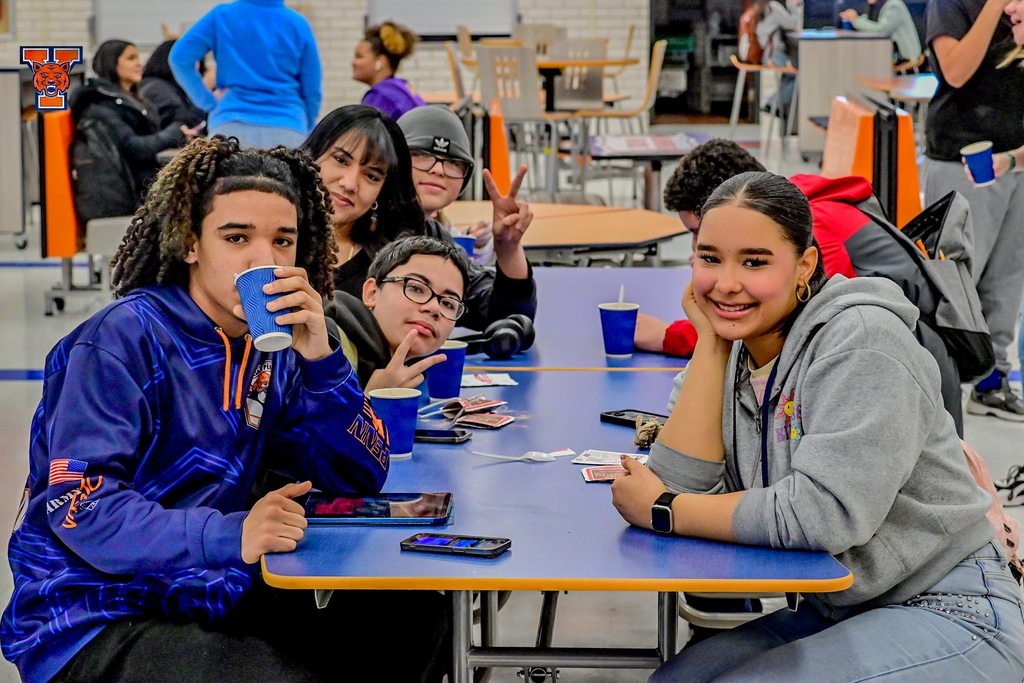 A group of five students are sitting at a table with cups on it in a school cafeteria. One of the students is drinking from the cup. Other people can be seen in the background.