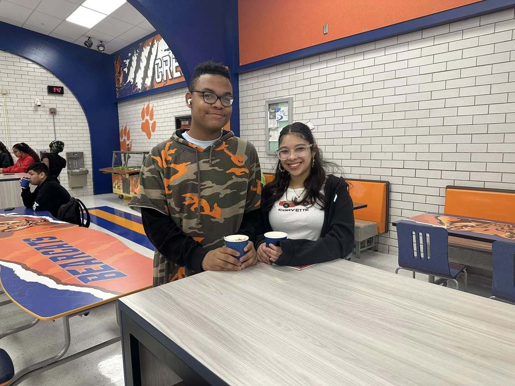 Two students are standing near a cafeteria table holding cups in a school cafeteria. The background features additional tables with the word "BEARCATS" on one of them. Other people can be seen in the background.