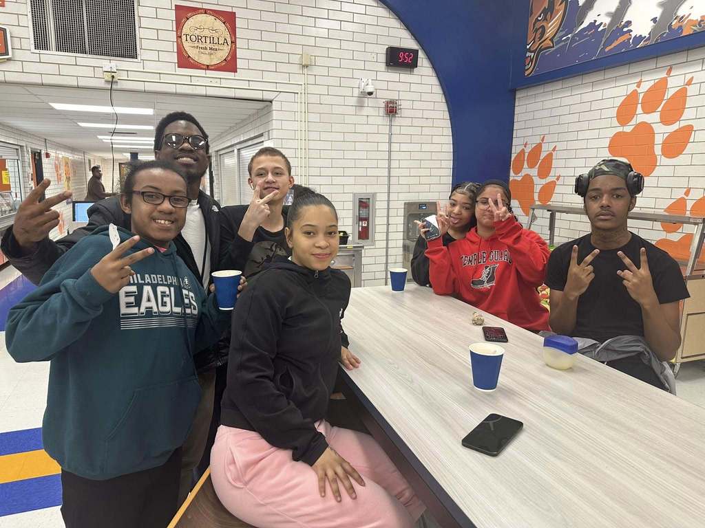 A district staff member and a group of six students are standing and sitting near a table that has cups on it in a school cafeteria. Other people can be seen in the background.