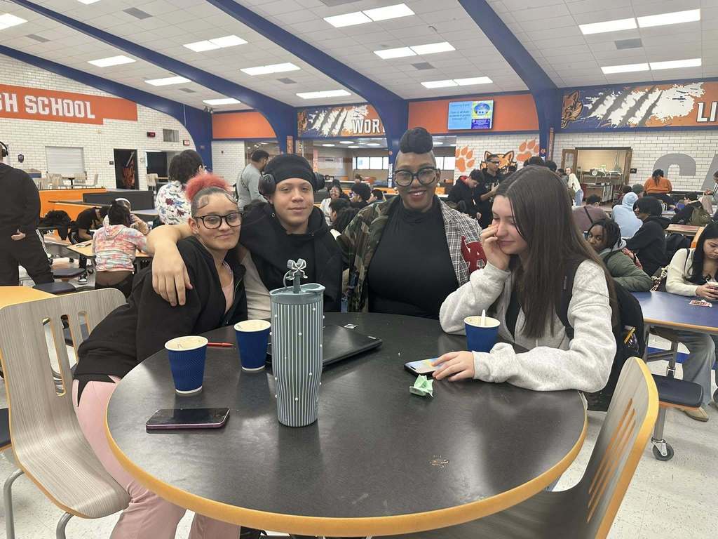 A William Penn Senior High School Administrator and a group of three students are sitting and standing around a table that has cups on it in a school cafeteria. Other people can be seen in the background.
