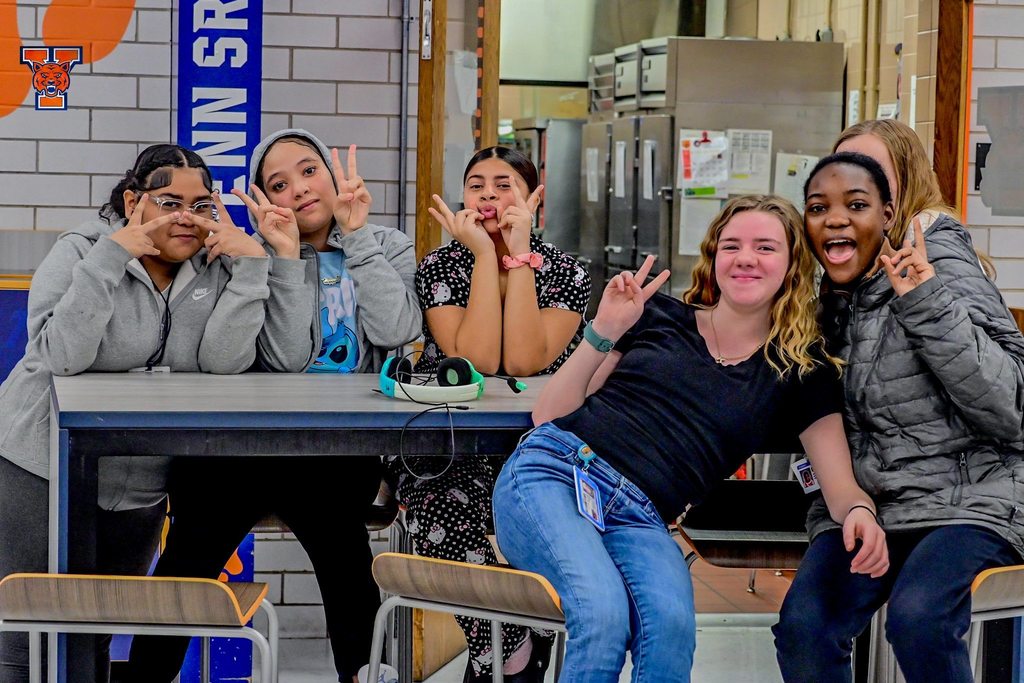A group of five students are sitting at a table in a school cafeteria holding up peace signs.