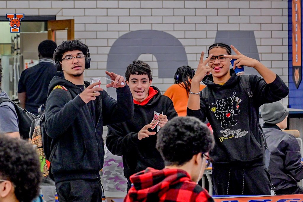 A group of three students are posing together in a school cafeteria, with two of them holding up peace signs. Other people can be seen nearby.