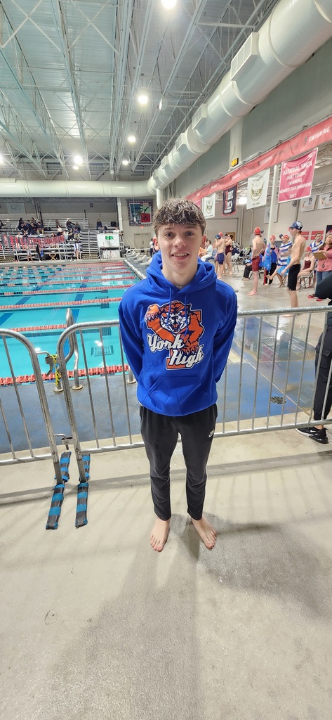 A William Penn Senior High School student is standing near a gate during an indoor swimming pool event. Other swimmers and people can be seen in the background.