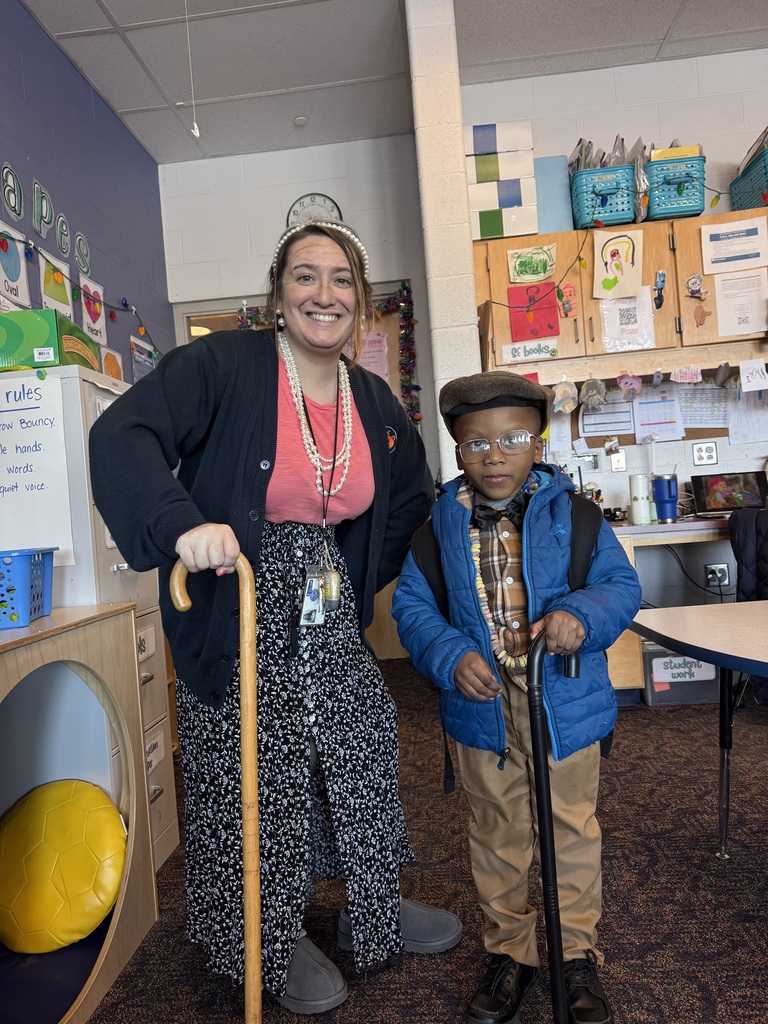 A teacher and a student, dressed as elderly people, are standing in a school classroom. Both of them are holding canes and smiling. 