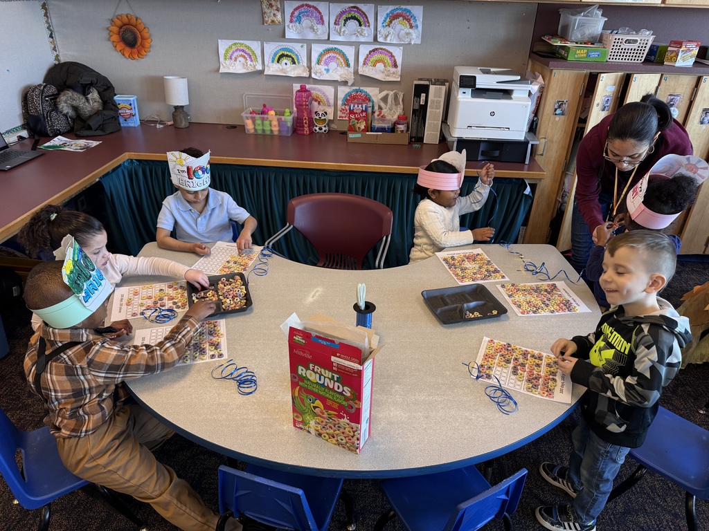 A group of six students wearing paper crowns sit are gathered around a table in a school classroom doing crafts using cereal. A teacher can be seen assisting one of the students. Art and educational supplies can be seen in the classroom.
