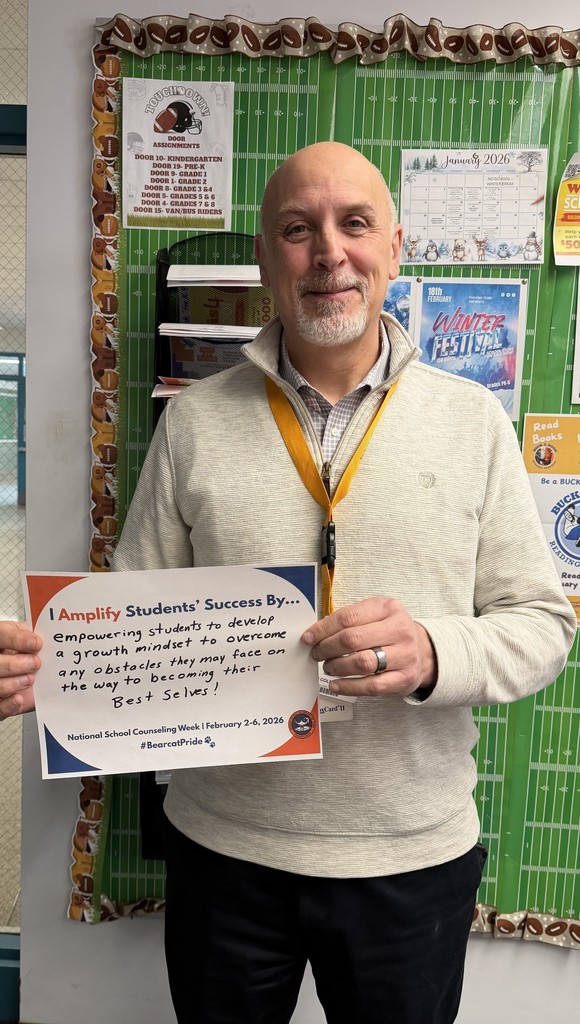 A district staff member is standing in front of a bulletin board, holding a sign that says "I Amplify Students' Success By empowering students to develop a growth mindset to overcome any obstacles they may face on the way to becoming their Best Selves!"