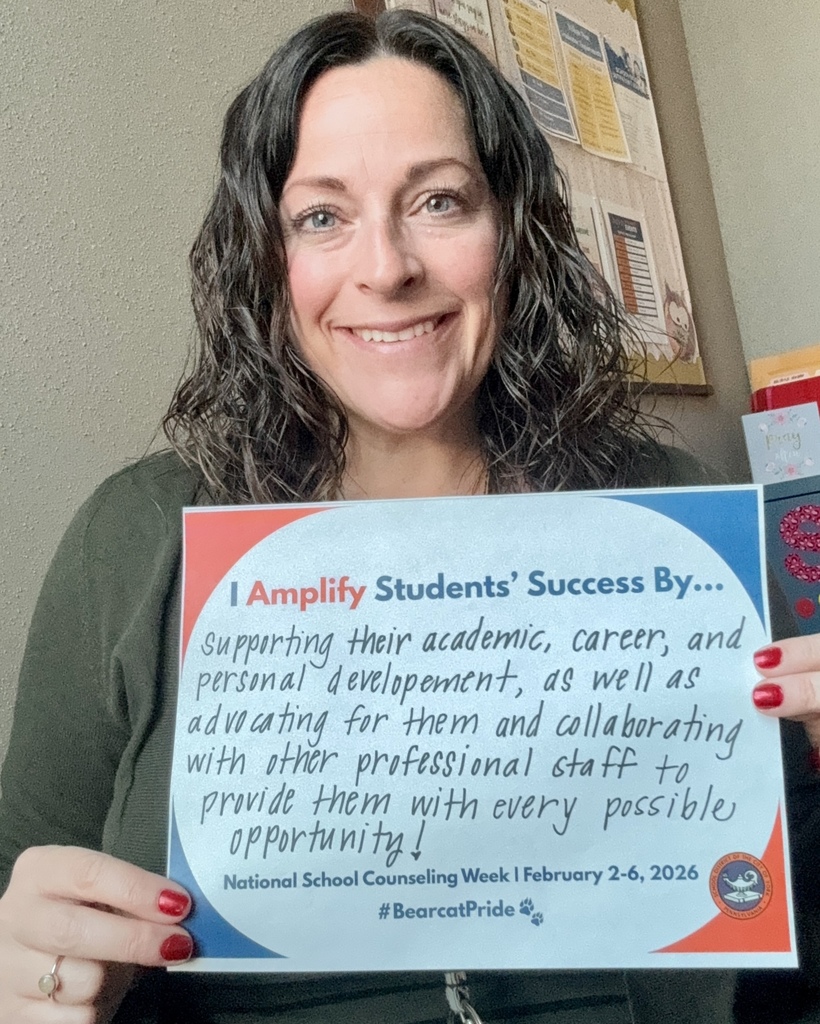 A district staff member is holding a sign that says "I Amplify Students' Success By supporting their academic, career, and personal development." An office bulletin board can be seen in the background.