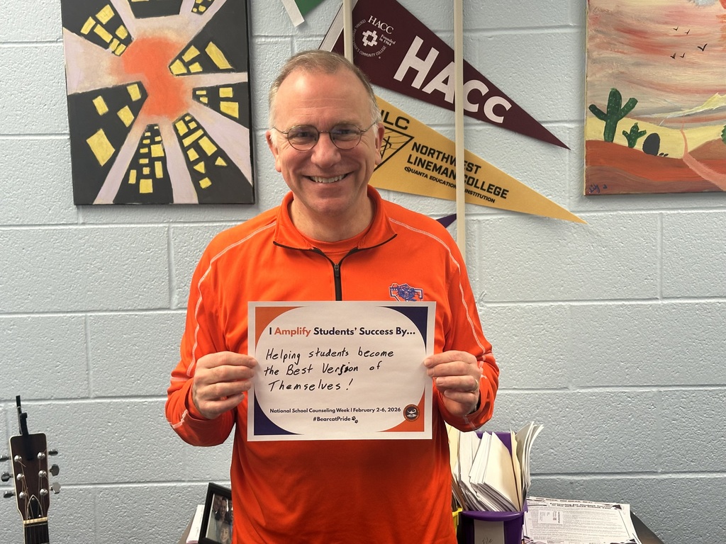 A district staff member is standing in front of a wall, holding a sign that says "I Amplify Students' Success By Helping students become the Best Version of Themselves!"