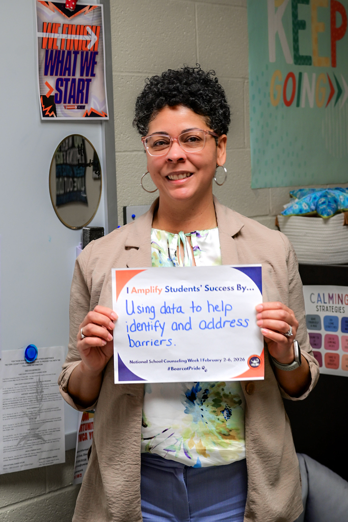 A district staff member is standing in a school classroom, holding a sign that says "I Amplify Students' Success By Using data to help identify and address barriers."