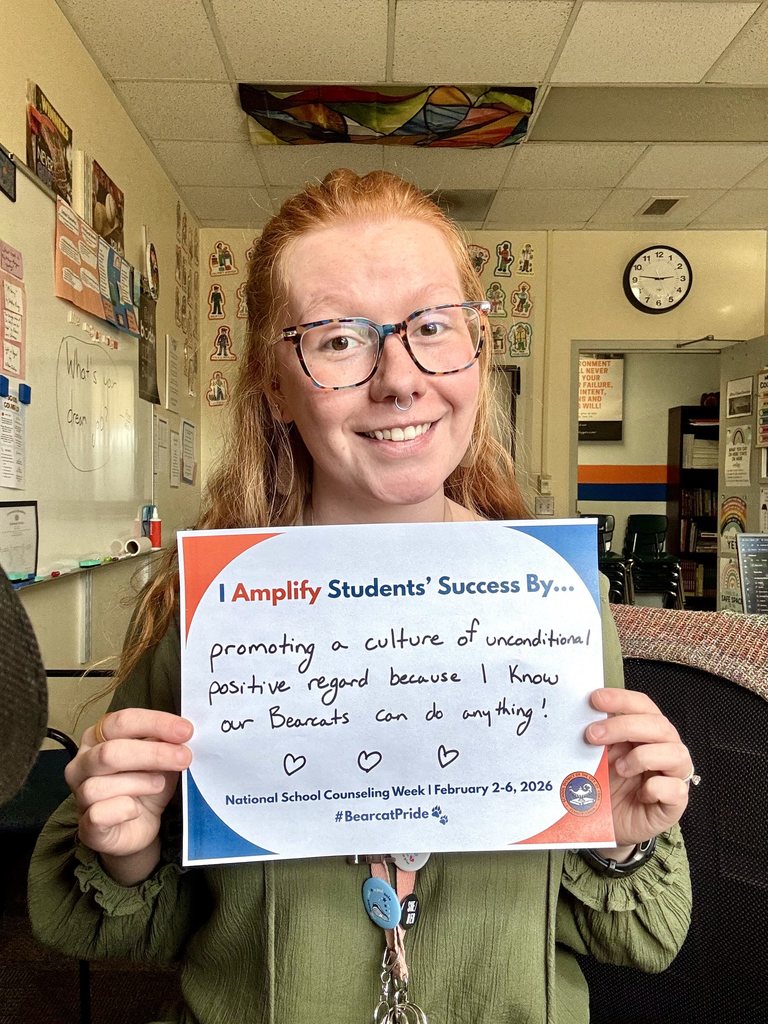 A district staff member is sitting in a school classroom, holding a sign that says "I Amplify Students' Success By promoting a culture of unconditional positive regard because I know our Bearcats can do anything!" with three hearts at the bottom.