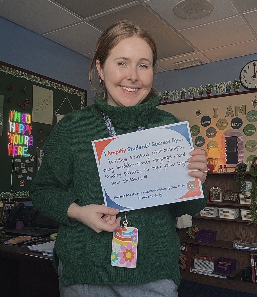 A district staff member is standing in a school classroom, holding a sign that says "I Amplify Students' Success By building trusting relationships, using strengths-based language, and showing patience as they grow beyond past mistakes." with a heart next to it.