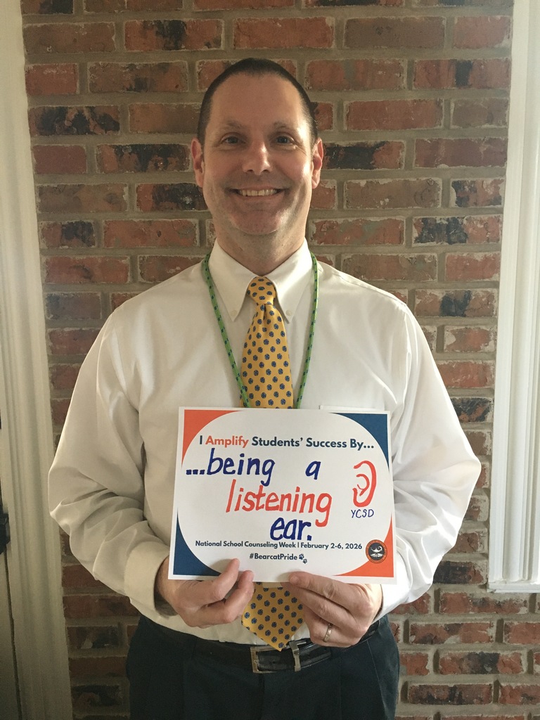 A district staff member is standing in front of a brick wall, holding a sign that says "I Amplify Students' Success By ...being a listening ear." A drawing of an ear can be seen next to the words.