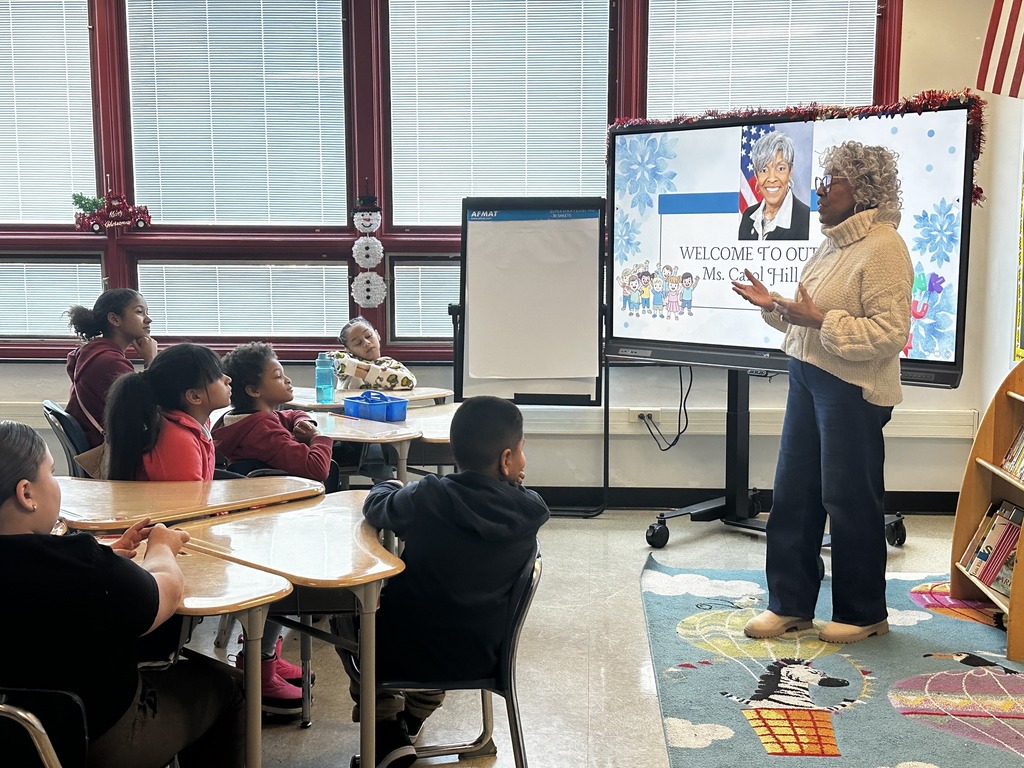 Representative Carol Hill-Evans is standing in front of a class of students, standing near a screen with text "Welcome to our class, Ms. Col. Hill" and a photo. The students are sitting attentively at desks facing Representative Carol Hill-Evans.