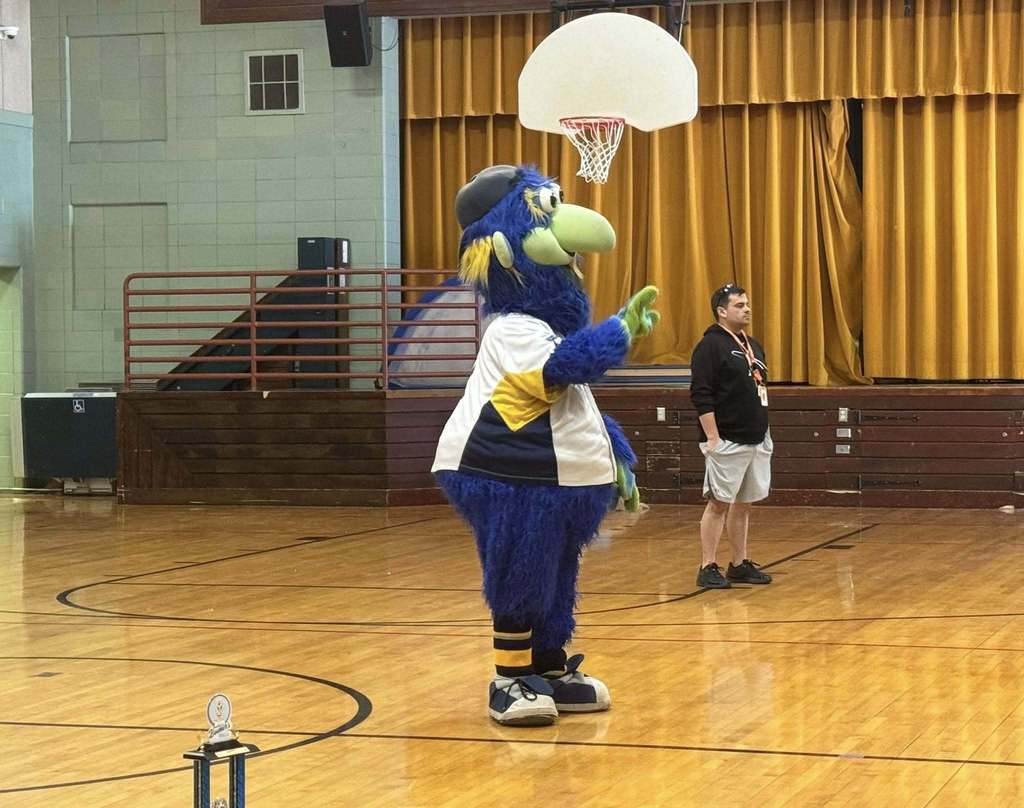 A person dressed as a blue and green bird mascot is waving one of their hands in the air while standing inside of a school gymnasium. A trophy can be seen nearby on the floor. A district staff member can also be seen standing nearby.