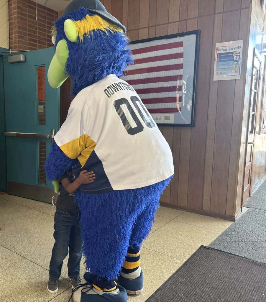 A person dressed as a blue and green bird mascot is hugging a small child in a school hallway. 