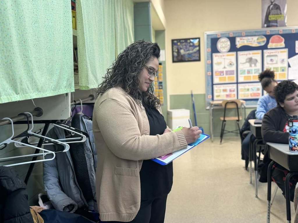 A district administrator is standing in a school classroom writing on a clipboard. Students can be seen sitting at desks nearby.