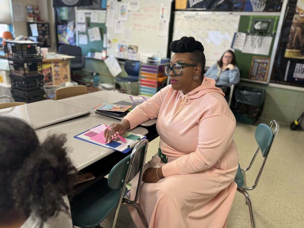 A district administrator is sitting at a desk in a school classroom, holding a pen over a clipboard. Another administrator can be seen nearby in the background.