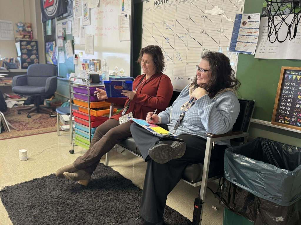 Two district administrators are sitting in a school classroom, one of them is looking down at a clipboard and the other has a pen in her hand.