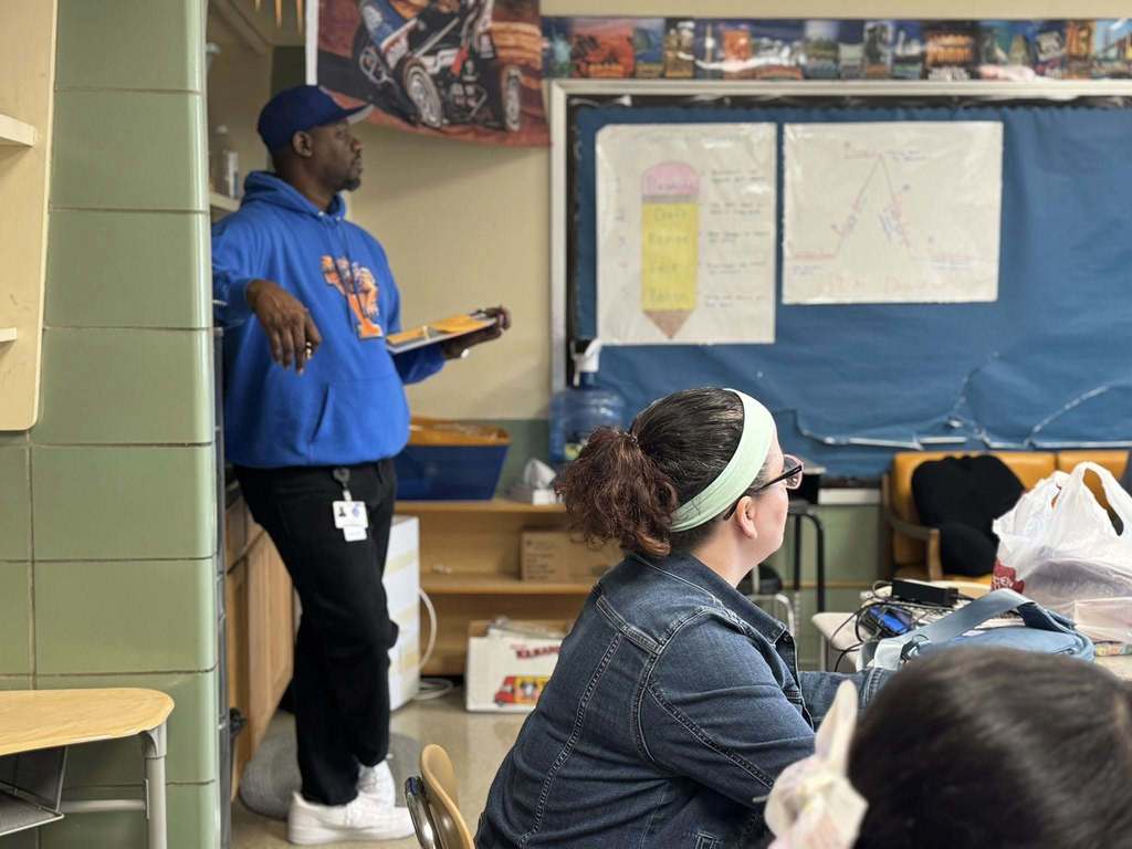 A district administrator is standing near a shelf in a school classroom with a clipboard, while another administrator is sitting at a desk.