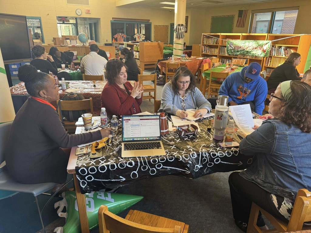 A group of five district administrators are sitting at a table in a school library, engaging in discussion. Other district administrators can be seen nearby in the background.