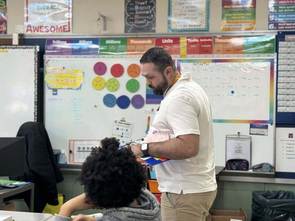 A district administrator is standing in a school classroom next to a student who is seated at a desk. 