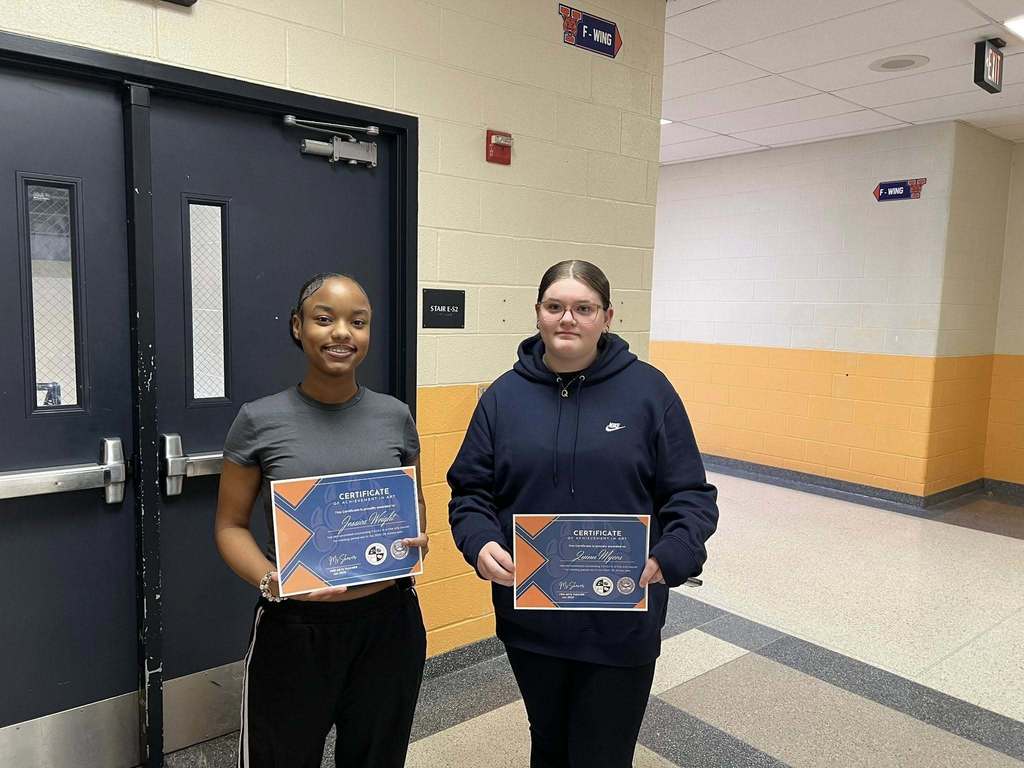 Two students are standing in a school hallway in front of double doors, holding certificates. 