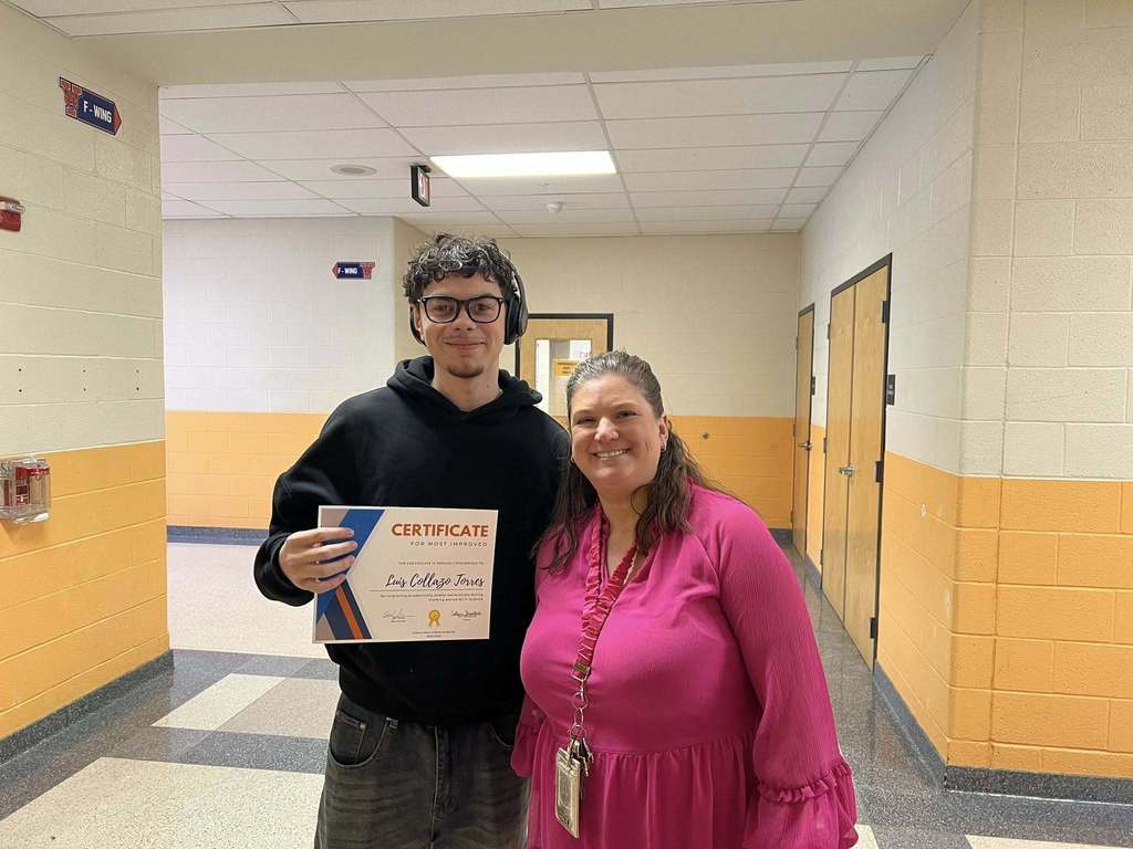 A district staff member and a student are standing in a school hallway. The student is holding a certificate in his hand. A door can be seen behind them in the background.