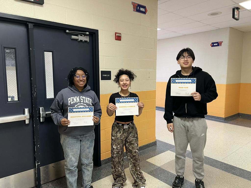 Three students are standing in a school hallway in front of double doors, holding certificates. 