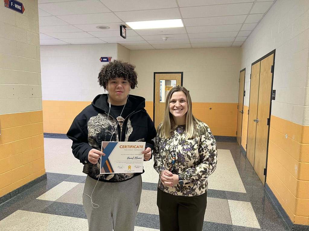 A district staff member and a student are standing in a school hallway. The student is holding a certificate in his hand. A door can be seen behind them in the background.