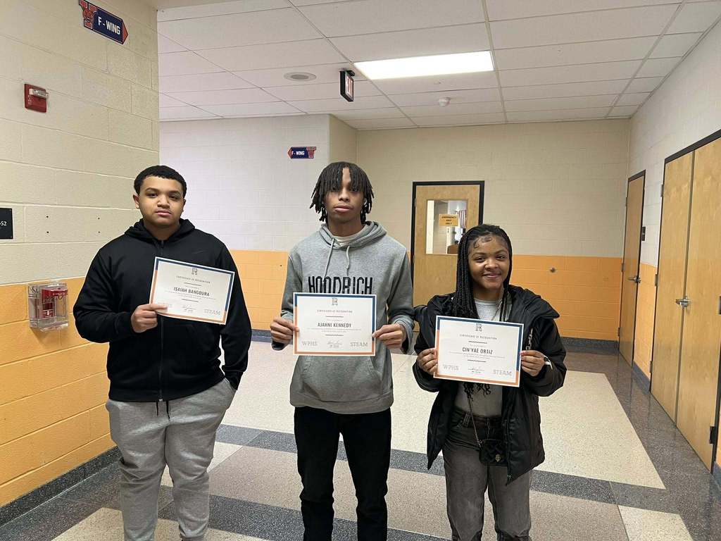 Three students are standing in a school hallway holding certificates. A door can be behind them in the background.