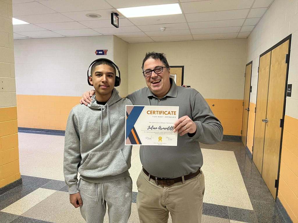 A district staff member and a student wearing headphones are standing in a school hallway. The district staff member is holding a certificate in one of his hands.