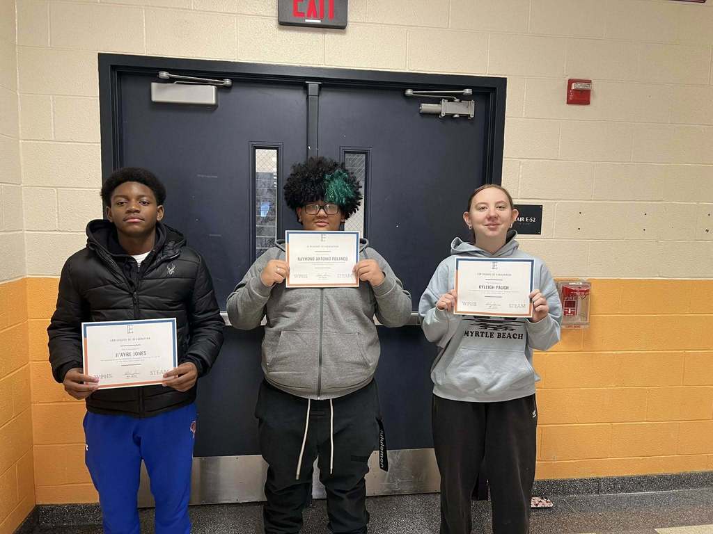 Three students are standing in a school hallway in front of double doors, holding certificates. 
