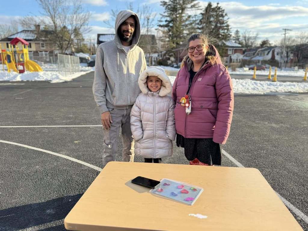 An adult man, a young girl, and a district staff member are standing outside in a parking lot behind a table that has a phone and colorful craft items on it.