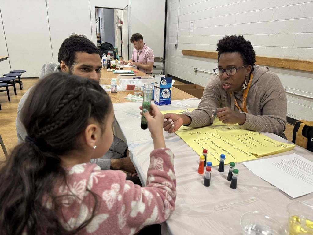 A group of three people, including a district staff member, a man and a young girl are sitting at a table in a school cafeteria. The district staff member is looking at the young girl who is holding a test tube in her hand. A man can be seen sitting to the left of the young girl. There is a poster and science materials that can be seen on the table. Another person can be seen in the background sitting at another table.