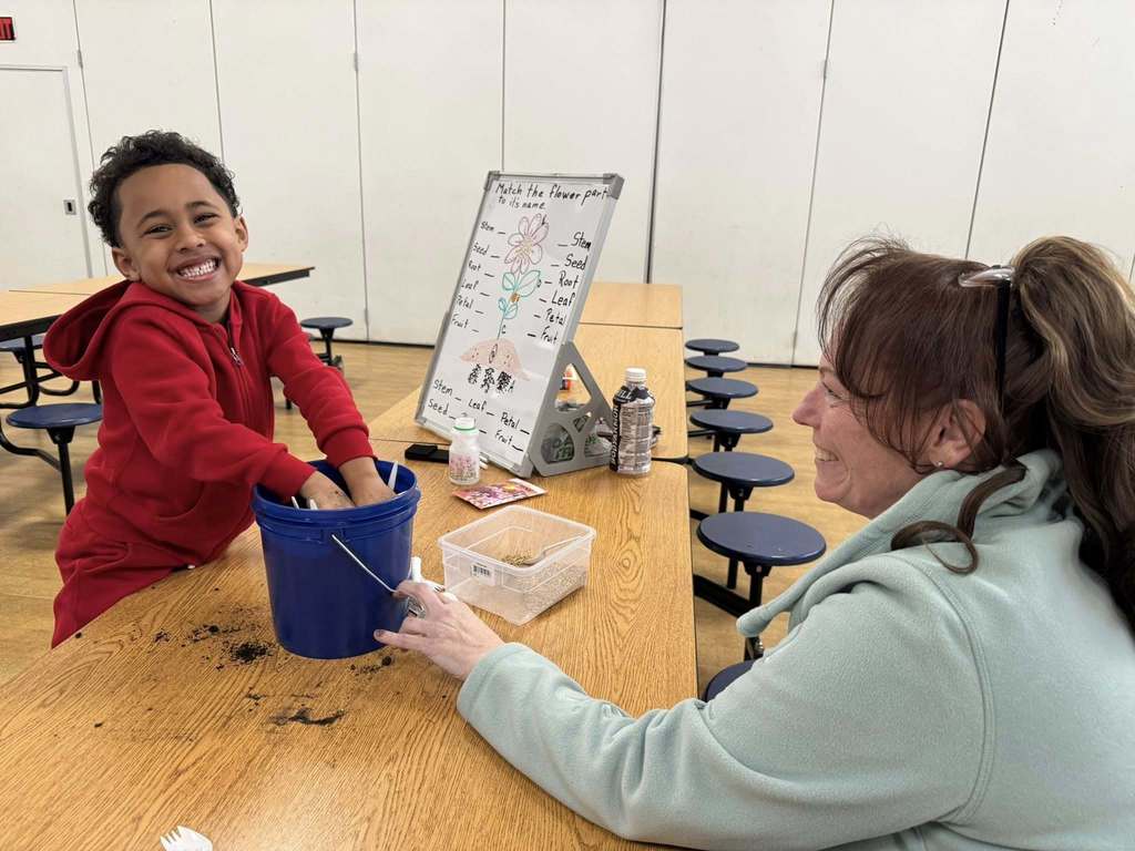 A young boy and a district staff member are sitting at a table in a school cafeteria. The young boy is joyfully digging into a blue pot of soil, while a district staff member is smiling at him. A board that has plant parts on it can be seen nearby on the table.