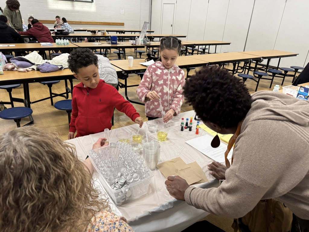 Two district staff members and two students are gathered around a table in a school cafeteria, engaging in a science experiment with pipettes and small containers.
