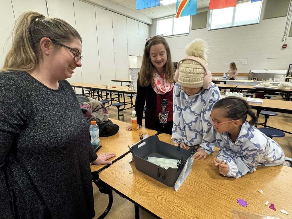 Two district staff members and two students are gathered around a table in a school cafeteria, examining a craft in a black bin. 