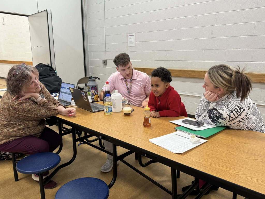 A young boy is sitting at a table in a school cafeteria, surrounded by three adults, including district administration. Laptops, drinks, and papers can be seen on the table. 