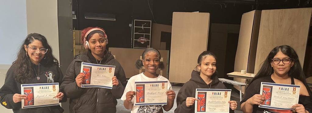  A group of five students are standing on a stage in a school auditorium, holding certificates in their hands. Wooden panels can be seen in the background.