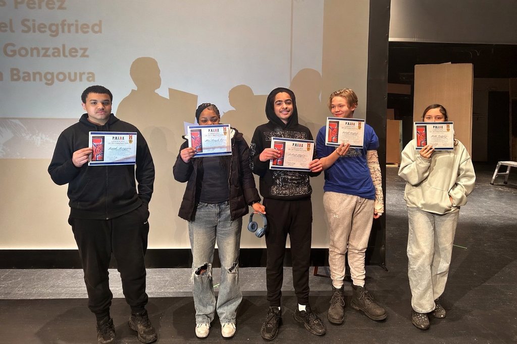A group of five students are standing on a stage in a school auditorium, holding certificates in their hands. A projector screen can be seen behind them.