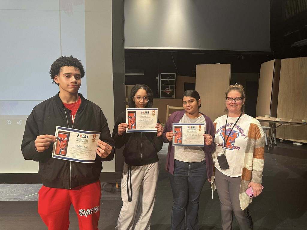A group of four students are standing on a stage in a school auditorium. A district staff member is located on the far right. Each of the students are holding certificates in their hands. A group of five students are standing on a stage in a school auditorium, holding certificates in their hands. A projector screen can be seen behind them.