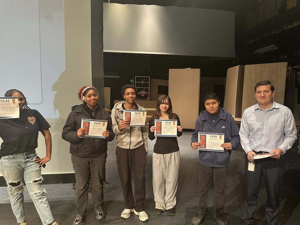 A group of five students are standing on a stage in a school auditorium, holding certificates in their hands. A district staff member can be seen standing to the far right. Wooden panels can be seen in the background.