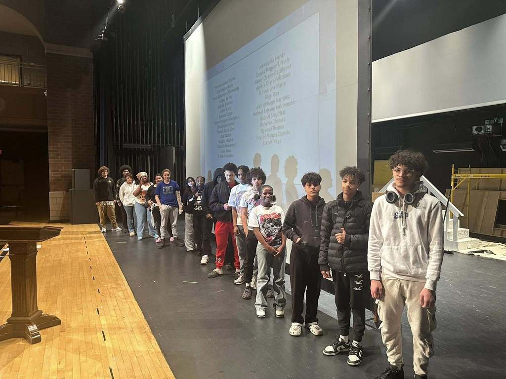 A group of students are standing in a line on a stage in a school auditorium. A projector screen with names on it can be seen behind them.
