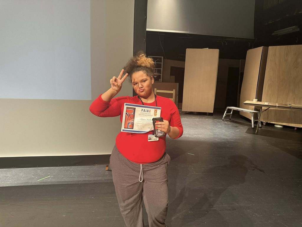A student is standing on a stage in a school auditorium, holding a certificate and a phone in her hand. A projector screen can be seen behind them.