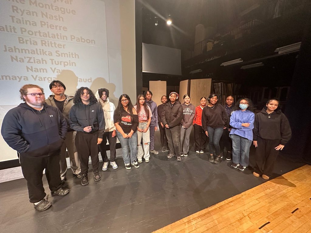 A group of students are standing in a line on a stage in a school auditorium. A projector screen with names on it can be seen behind them.