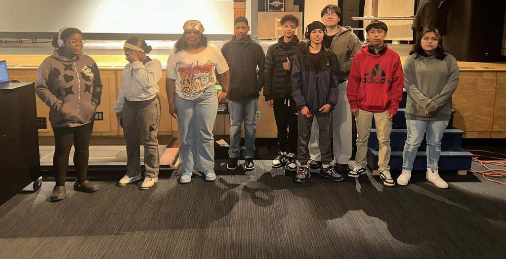 A group of nine students are standing side by side in a school auditorium. Some of the students are looking at the camera, while other students are looking away from the camera.