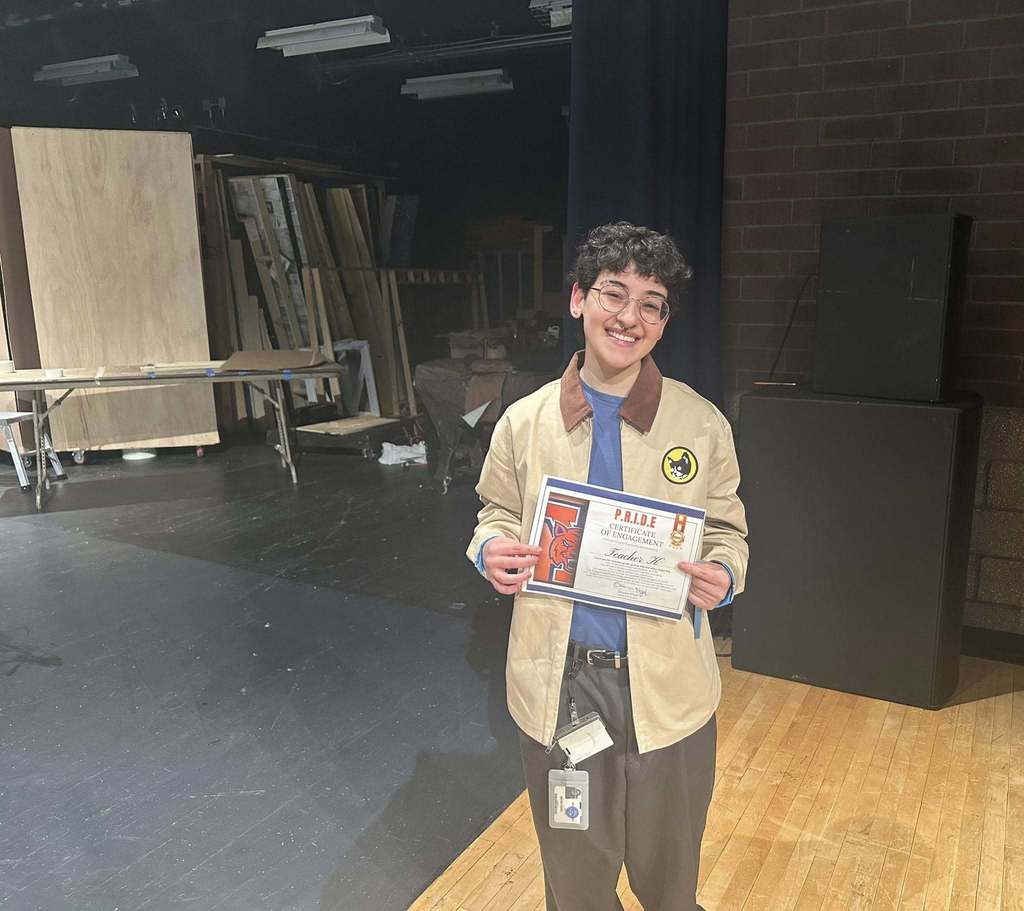 A district staff member is standing on a stage in a school auditorium, holding a certificate. Wooden panels and tables can be seen in the background.